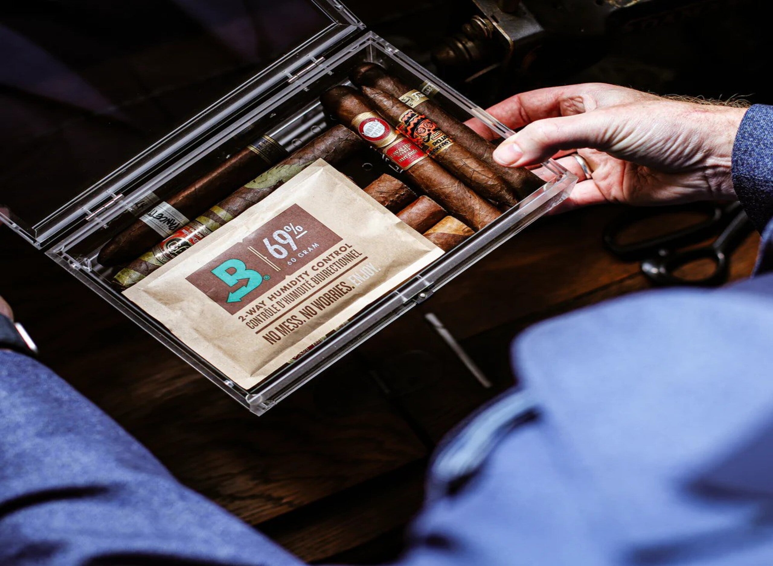 Wooden box of cigars on a table, surrounded by loose tobacco, bottles, and a potted plant; warm lighting